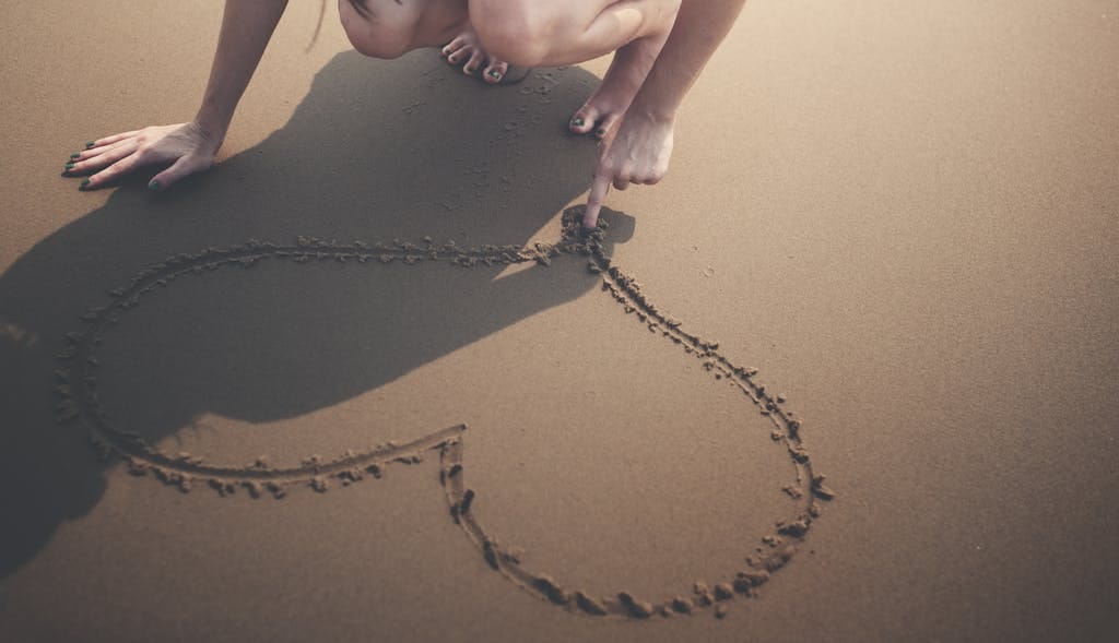 A woman drawing a heart shape in the sand with her finger on a sunny beach day.