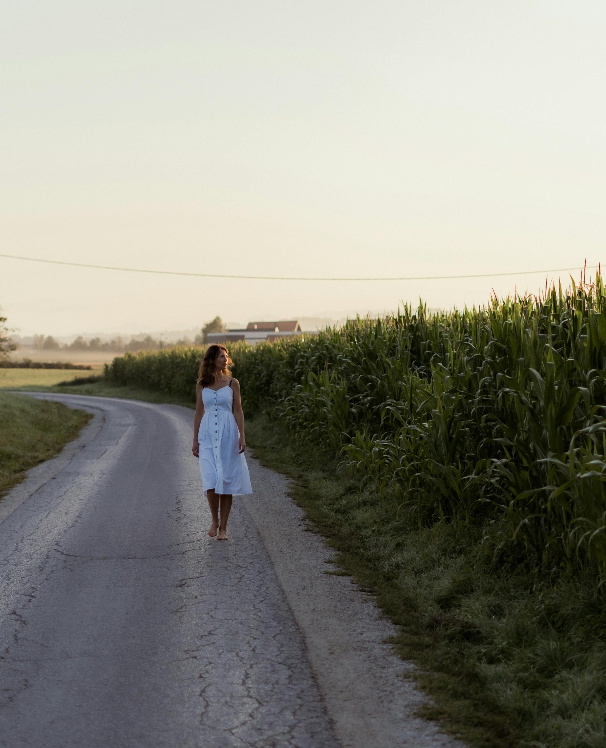 A woman in a white dress walking along a rural road surrounded by cornfields at sunset.